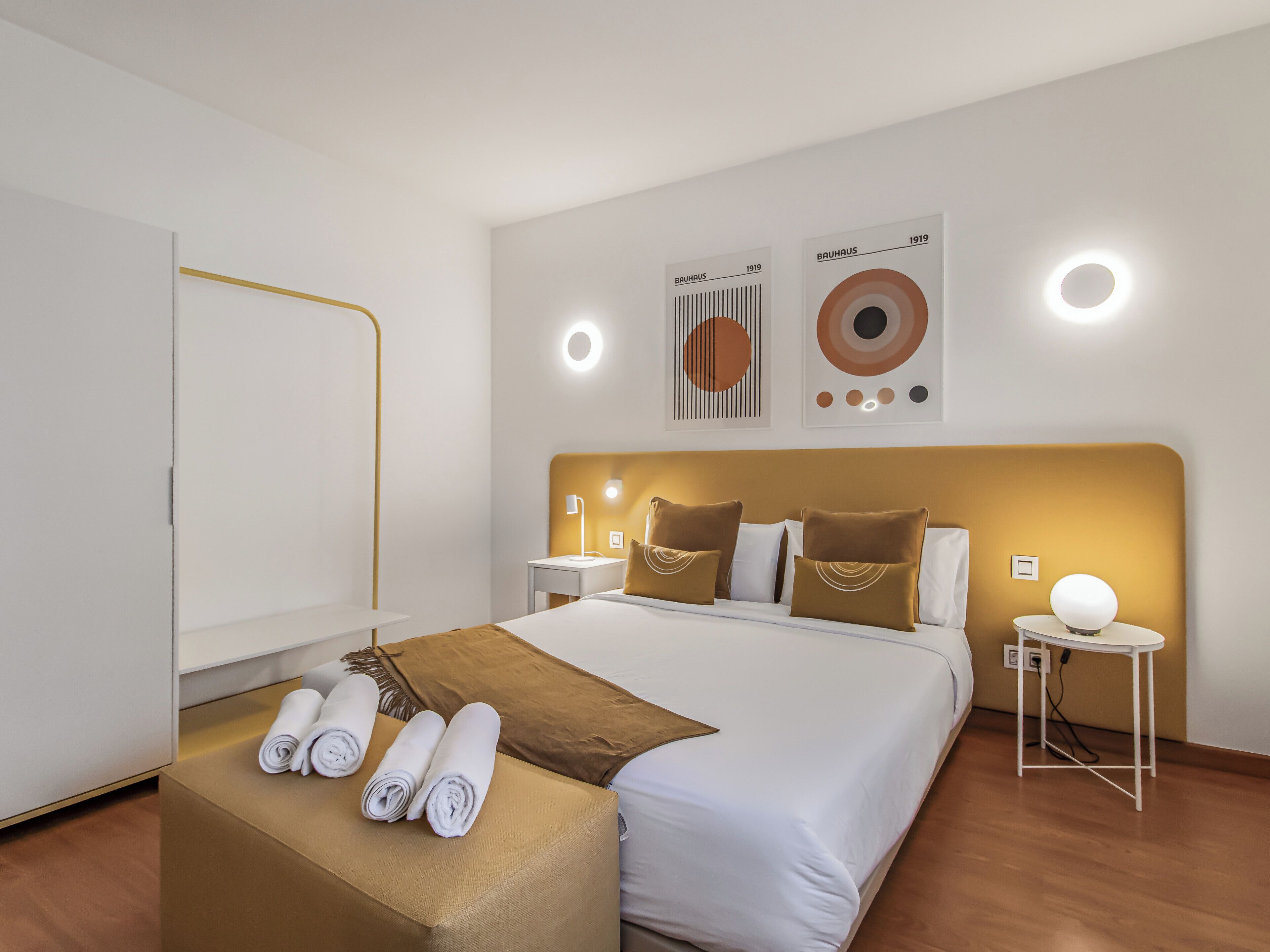 A modern hotel room with a large bed, a bedside table, a nightstand, and a wall-mounted light fixture.