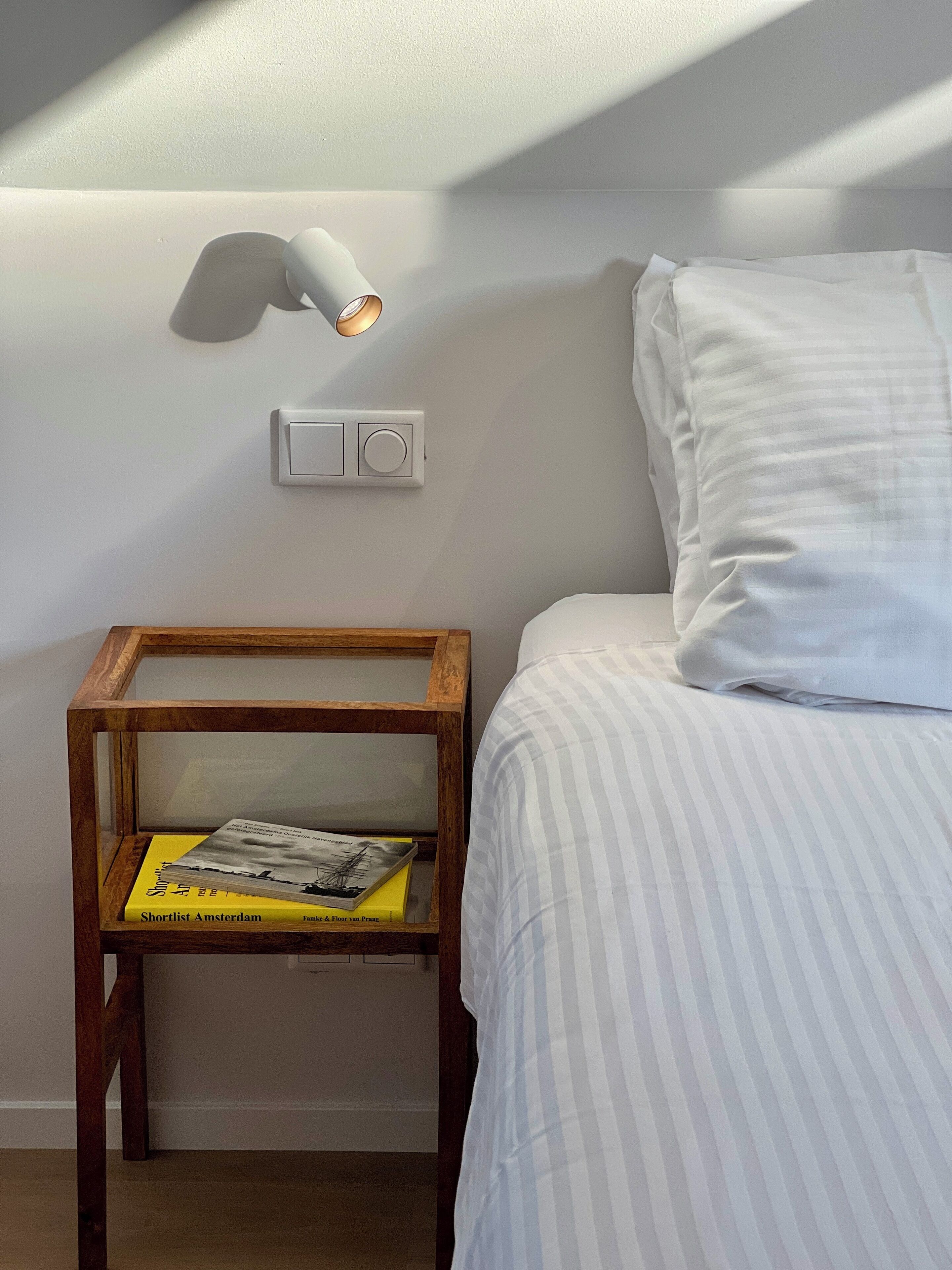 Ein ordentlich bezogenes Bett mit weißer Streifensteppdecke, einer Nachttischlampe und einem Holfnachtkästchen mit einer Zeitschrift.