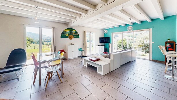 A spacious living area with a white sofa, a glass dining table with chairs, and a TV mounted on the wall. - Belle Maison vue montagnes (Bidarray)