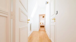 Een hal met witte muren en een houten vloer, die leidt naar een slaapkamer met een bed en nachtkastje.