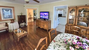 A living room with a ceiling fan, a television, a dining table with floral tablecloth, and a kitchen area visible through an open doorway. - Magnolia Vacation Rental  w/ Porch & Pond (Senatobia)