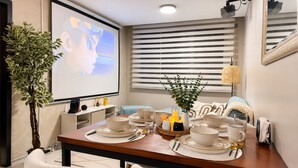 Un coin repas avec une table dressée pour quatre personnes, un écran de projection, une plante et une fenêtre avec des stores.