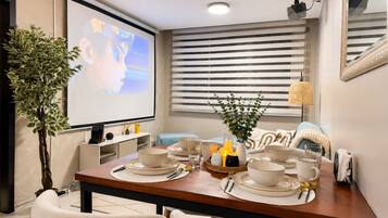 A dining area with a table set for four, a projector screen, a plant, and a window with blinds.