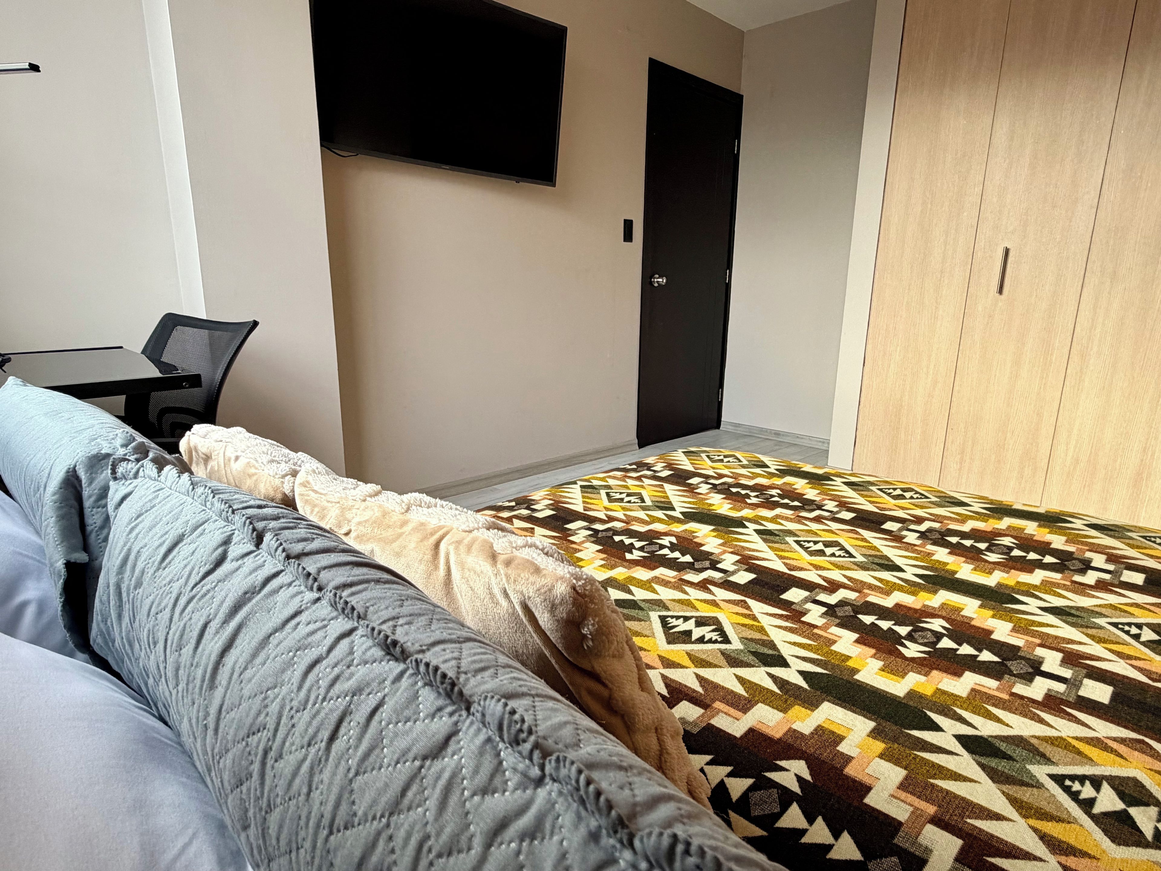 Ein Schlafzimmer mit einem Bett, einem Schreibtisch, einem Stuhl, einem an der Wand montierten Fernseher und einem Kleiderschrank.