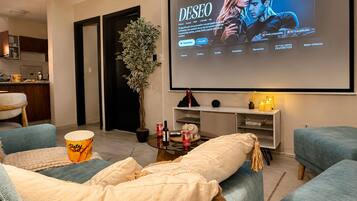 A living room with a large screen displaying a movie, comfortable seating, a small table with snacks, and a potted plant.