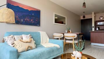 Una sala de estar con un sofá azul, una mesa de centro de vidrio y un cuadro de un paisaje urbano en la pared.