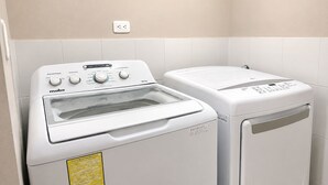 Une machine à laver et un sèche-linge à chargement frontal, placés côte à côte dans une buanderie aux murs recouverts de carreaux blancs, avec une prise de courant au-dessus des appareils.