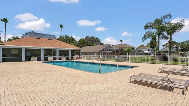 Piscina con sillones de descanso y una zona techada, rodeada de una cerca y palmeras.