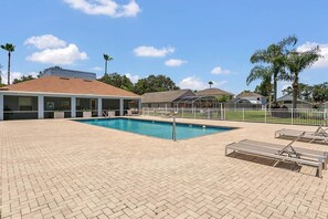 Piscina con sillones de descanso y una zona techada, rodeada de una cerca y palmeras.