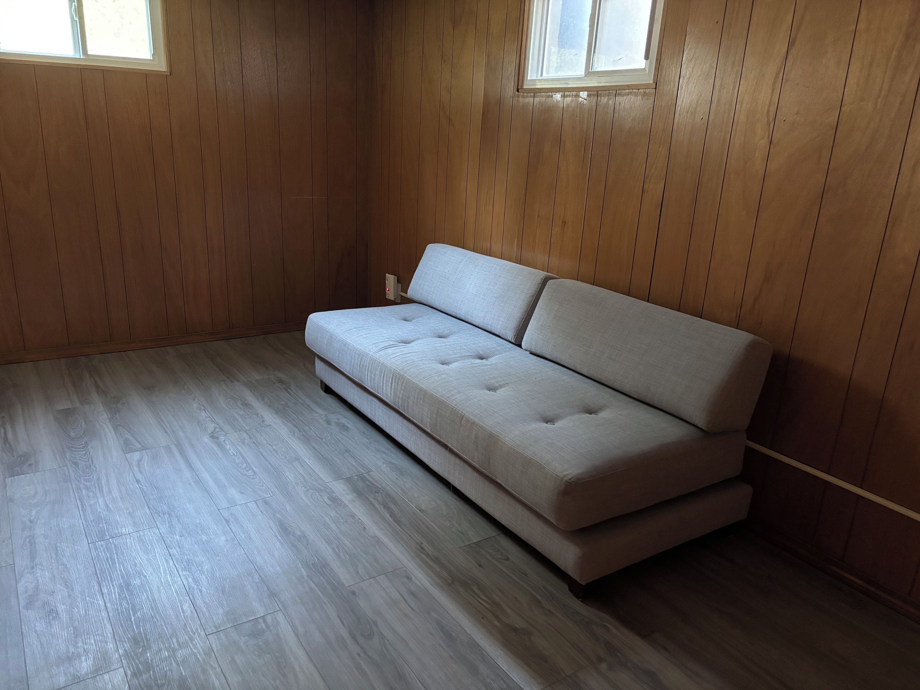 Une pièce avec un canapé de couleur claire, des murs en bois, une fenêtre et un plancher en bois.