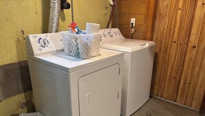 Un área de lavandería con dos lavadoras blancas, un cesto de basura blanco y una pared de madera.