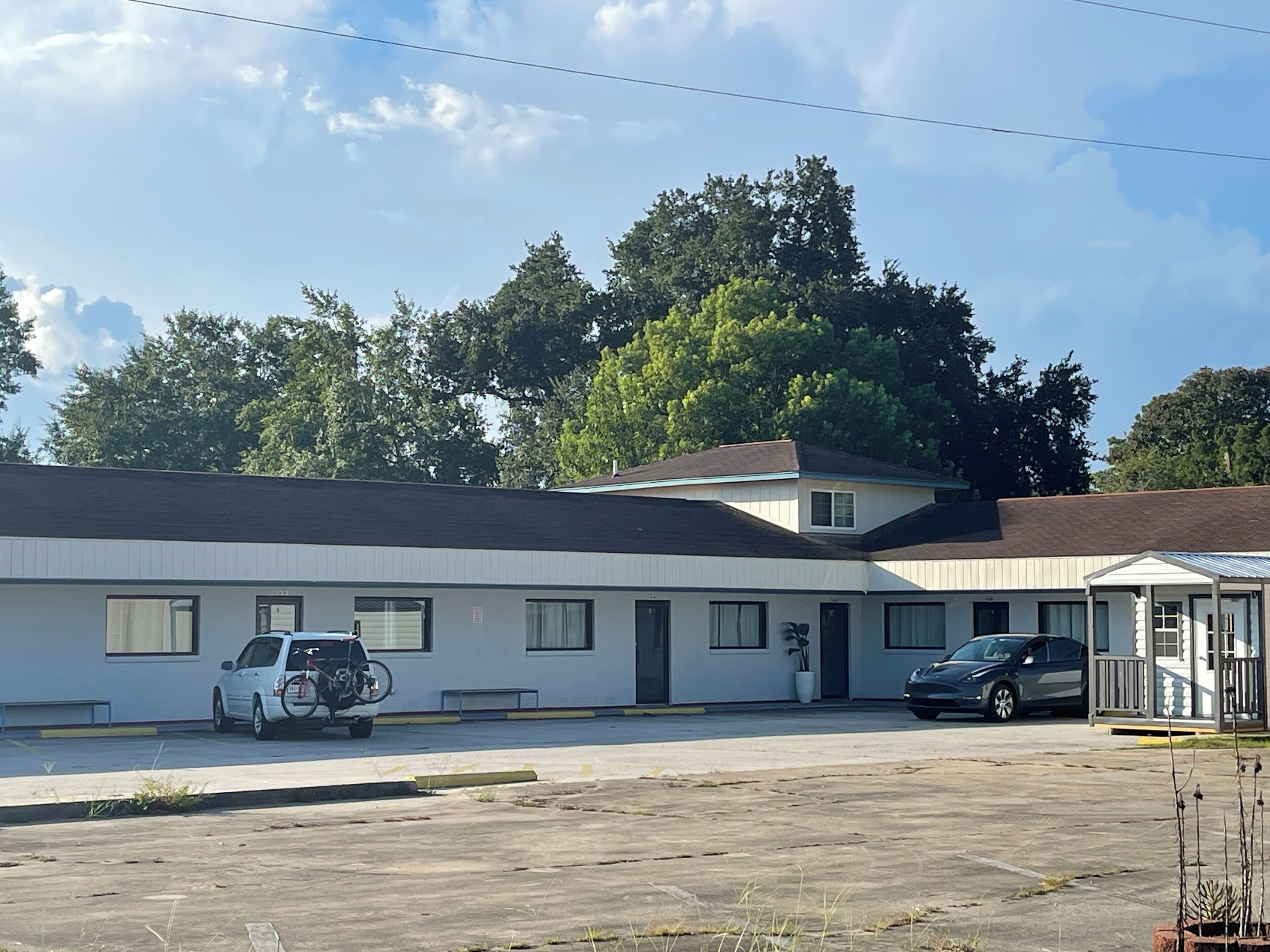 Ein eingeschossiges Motel mit weißer Fassade und dunklem Dach. Auf dem Parkplatz stehen zwei Autos, eines weiß und eines dunkel.