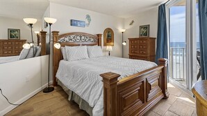 A bedroom with a large bed, wooden headboard, bedside lamps, a dresser, and a view of the ocean through a sliding glass door.