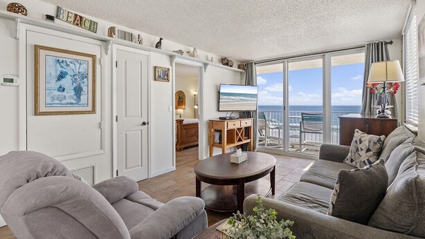 A spacious living room with a large window offering a view of the beach, a television, and a cozy seating area.