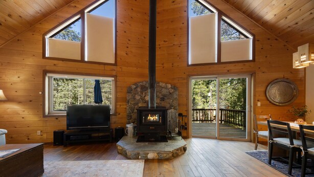A cozy cabin interior with a stone fireplace, wooden walls, large windows, and a dining area.