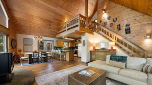 A spacious living area with a high ceiling, wooden beams, a staircase, a kitchen, and a dining area.