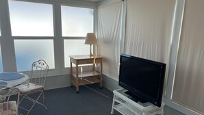 Une pièce avec un téléviseur posé sur un meuble blanc, une petite table avec une lampe, un fauteuil en osier et une vue sur la mer par de grandes fenêtres.