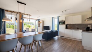 Une cuisine moderne avec des armoires blanches, un plan de travail en bois et un four encastré. On y trouve un coin repas avec une table et des chaises en bois, ainsi qu’une grande fenêtre offrant une vue sur l’extérieur.