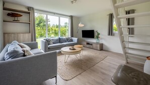 A modern living room with a sofa, coffee table, and TV. There is a staircase to the right and a window with a view of greenery outside. - Gorgeous home in Kamperland with WiFi (Kamperland)