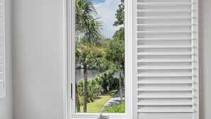 A window with white shutters, a view of a lush garden with palm trees and a pathway, and a body of water in the background. - Windswept 2BD Condo, Oceanside Kiawah, Steps to Beach & Park, Lagoon Views! (Kiawah Island)