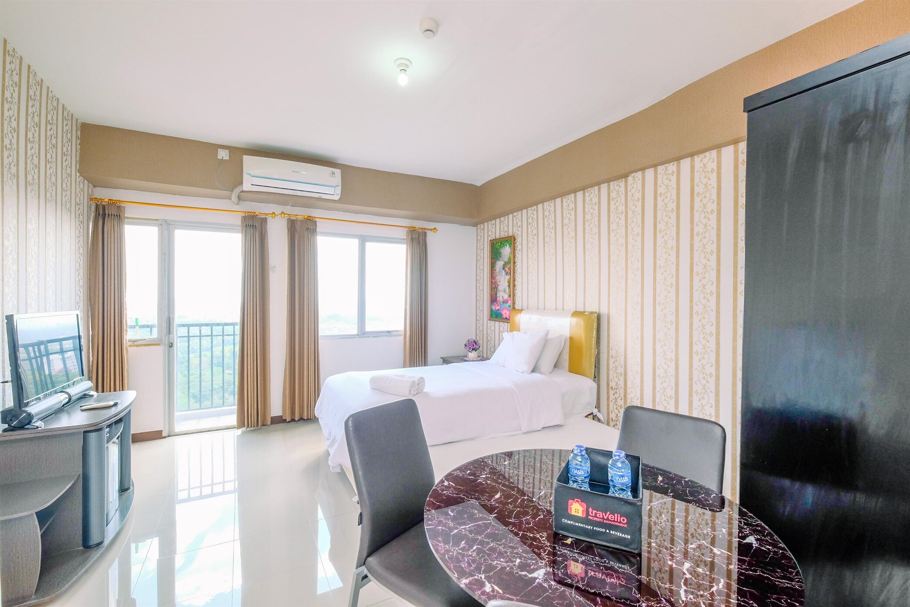 A hotel room with a bed, a desk with a TV, a dining table, and a large window.