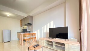 Egy modern nappali, ahol van egy televízió, fából készült bútorok, és a háttérben egy konyha látható.