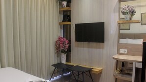 Habitación de hotel con cama, televisor montado en la pared, escritorio pequeño con silla, florero con flores y zapatero.