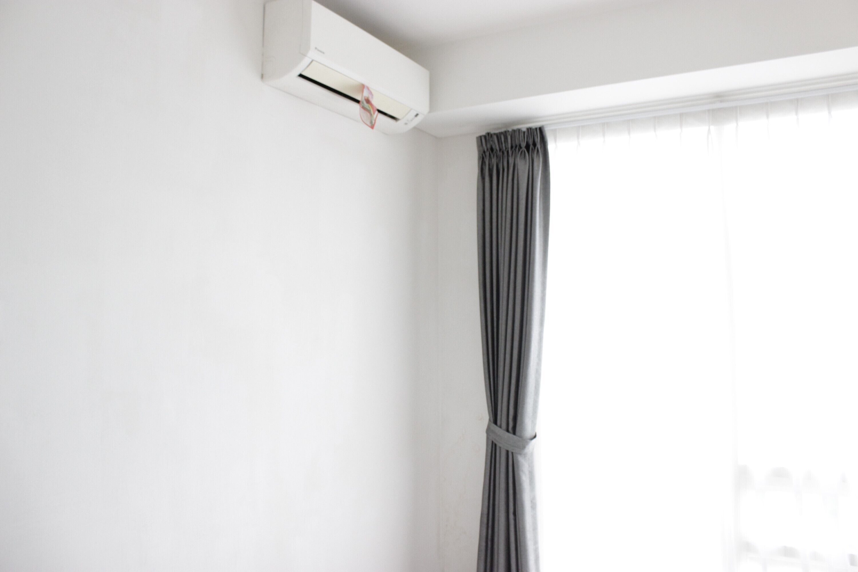A room with a white wall, a window with grey curtains, and an air conditioning unit mounted on the wall.