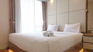 Egy gondosan megterített ágy fehér ágyneművel és szépen összehajtogatott törölközőkkel, egy fafejű ágykeret, és egy éjjeliszekrény, rajta egy lámpával.