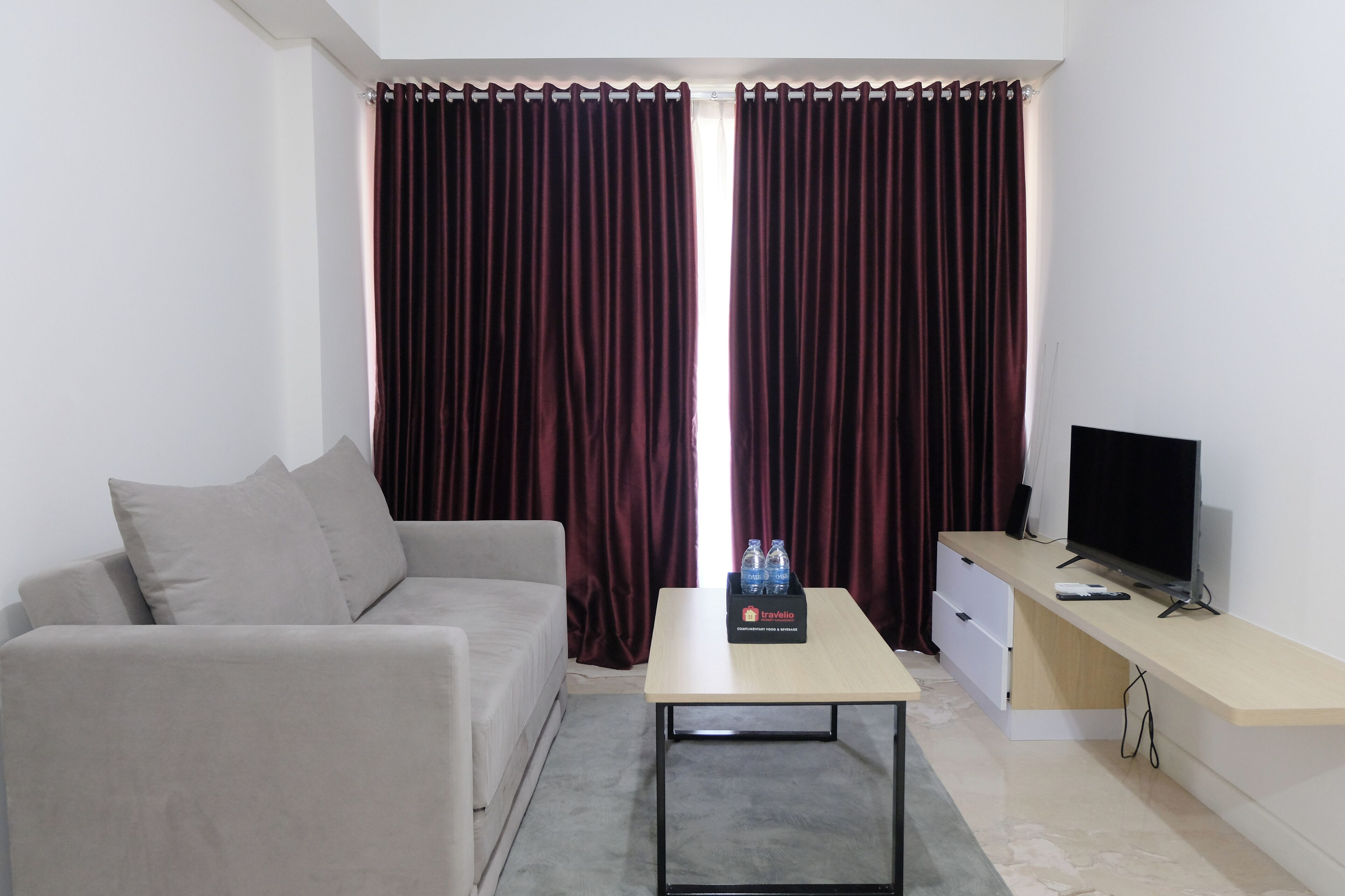 A modern living room with a grey sofa, a coffee table, a flat-screen TV, and a minibar with water bottles.