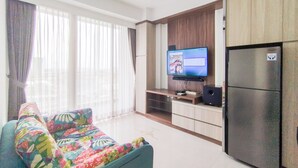 A living room with a sofa, a flat-screen TV mounted on the wall, a refrigerator, and a window with curtains.