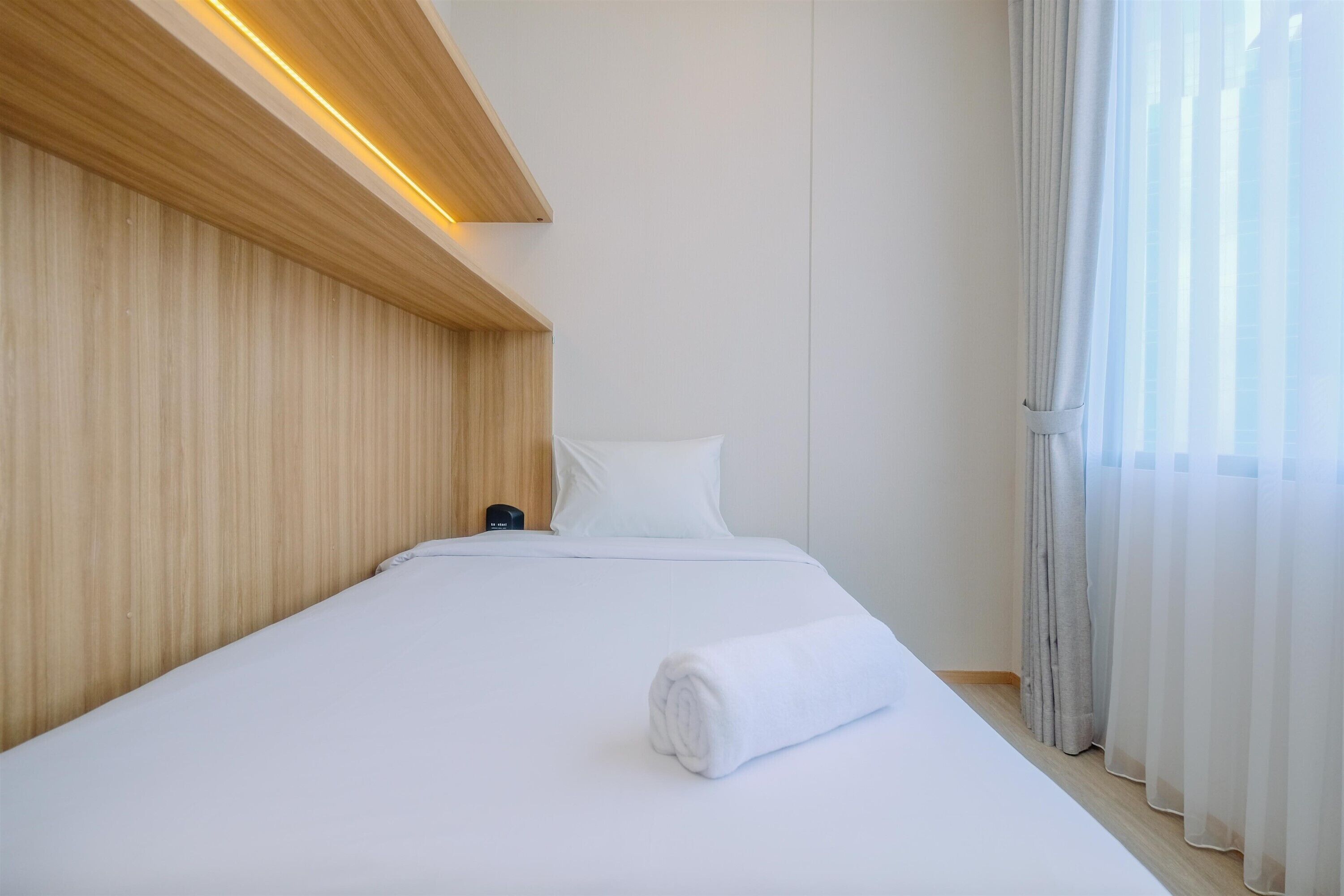 A hotel room with a neatly made bed, wooden headboard, and a window with sheer curtains.