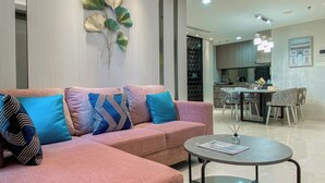 A modern living room with a pink sofa, blue and patterned cushions, a glass coffee table, and a decorative wall piece.