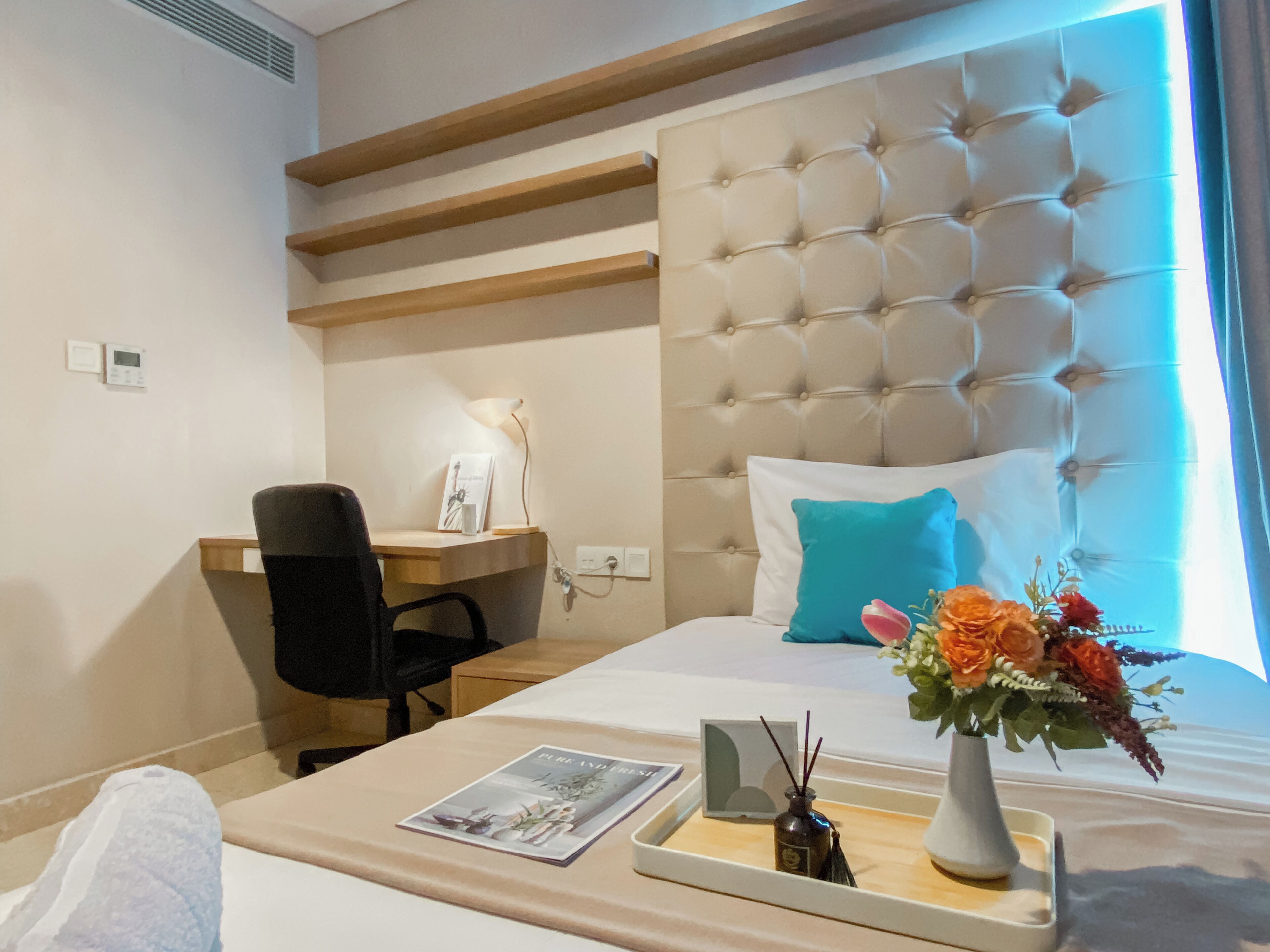 A hotel room with a tufted headboard, a desk with a chair, a bedside table with a vase of flowers, and a bed with a blue pillow.