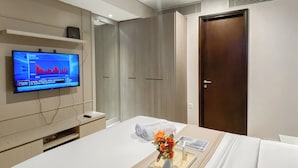 A hotel room with a bed, a TV displaying a news channel, a wooden door, and a bedside table with a vase of flowers.