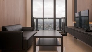 A modern living room with a large window, a sofa, a coffee table, and a TV on a wooden cabinet. - Comfy And Modern Look 2Br At Yukata Suites Apartment (South Tangerang)
