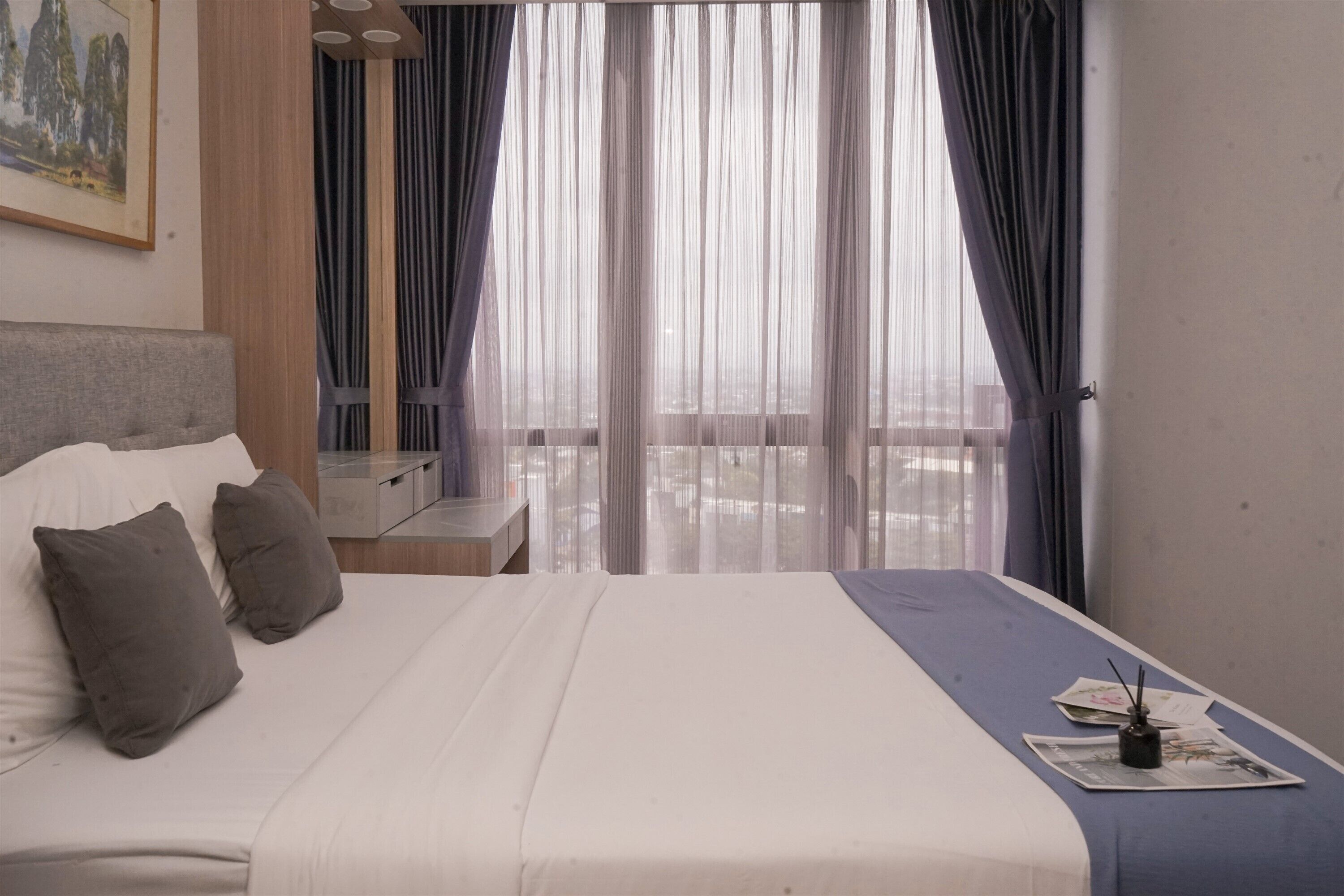 A hotel room with a bed, pillows, a nightstand, a small table, and a view of the outdoors through sheer curtains.