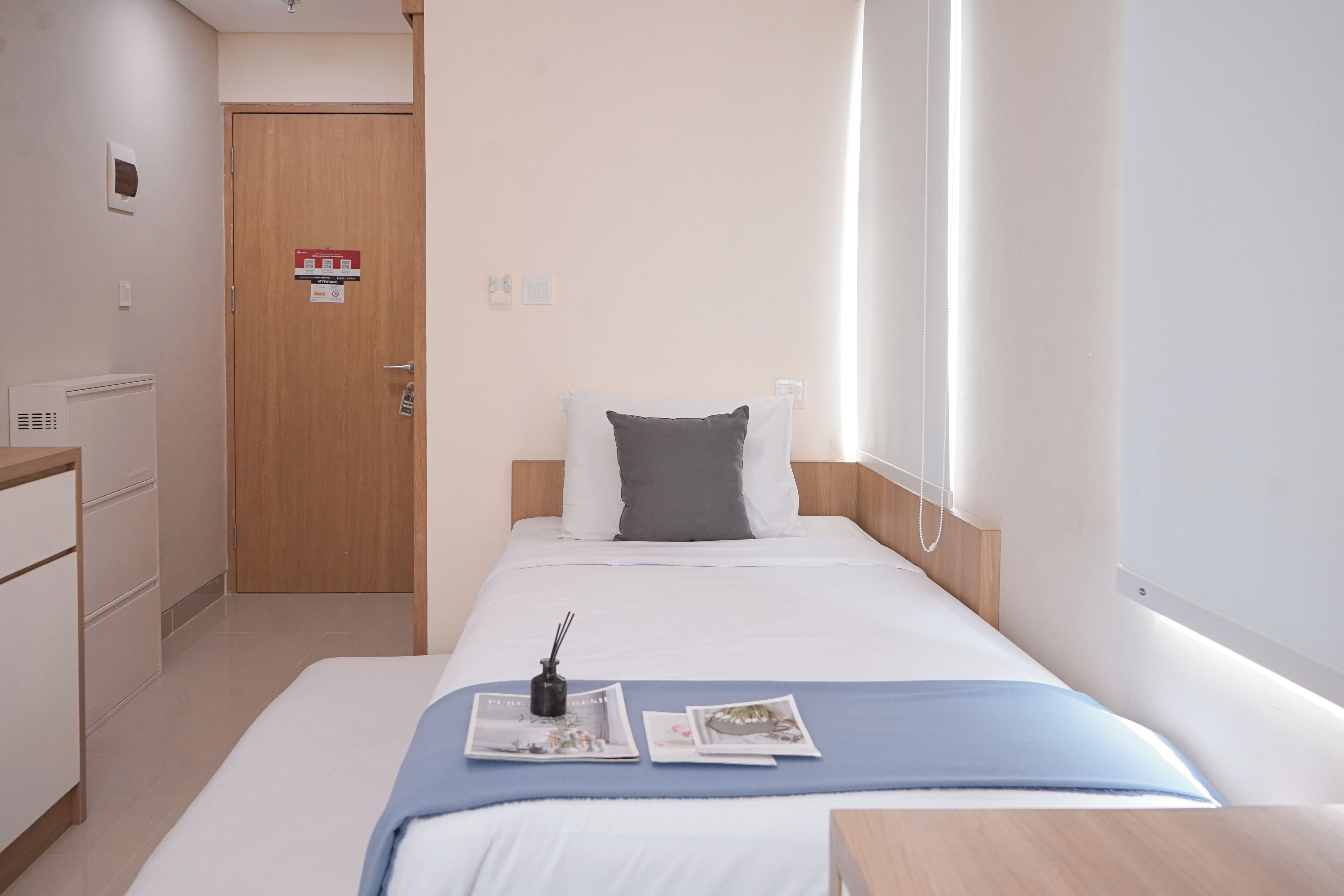 A hotel room with a bed, a bedside table, a nightstand, a wooden door, and a wall-mounted light fixture.