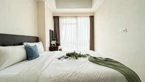 A neatly made bed with white and blue pillows, a green throw, and a magazine on the bed. There is a nightstand with a lamp and a potted plant. The room has a large window with sheer curtains and a view of buildings outside. - Comfort Living 2Br At Menteng Park Apartment (Jakarta)