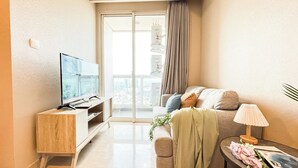 A modern living room with a flat-screen TV on a wooden stand, a sofa with cushions, a small table with a lamp and flowers, and a sliding glass door leading to a balcony. - Comfort Living 2Br At Menteng Park Apartment (Jakarta)