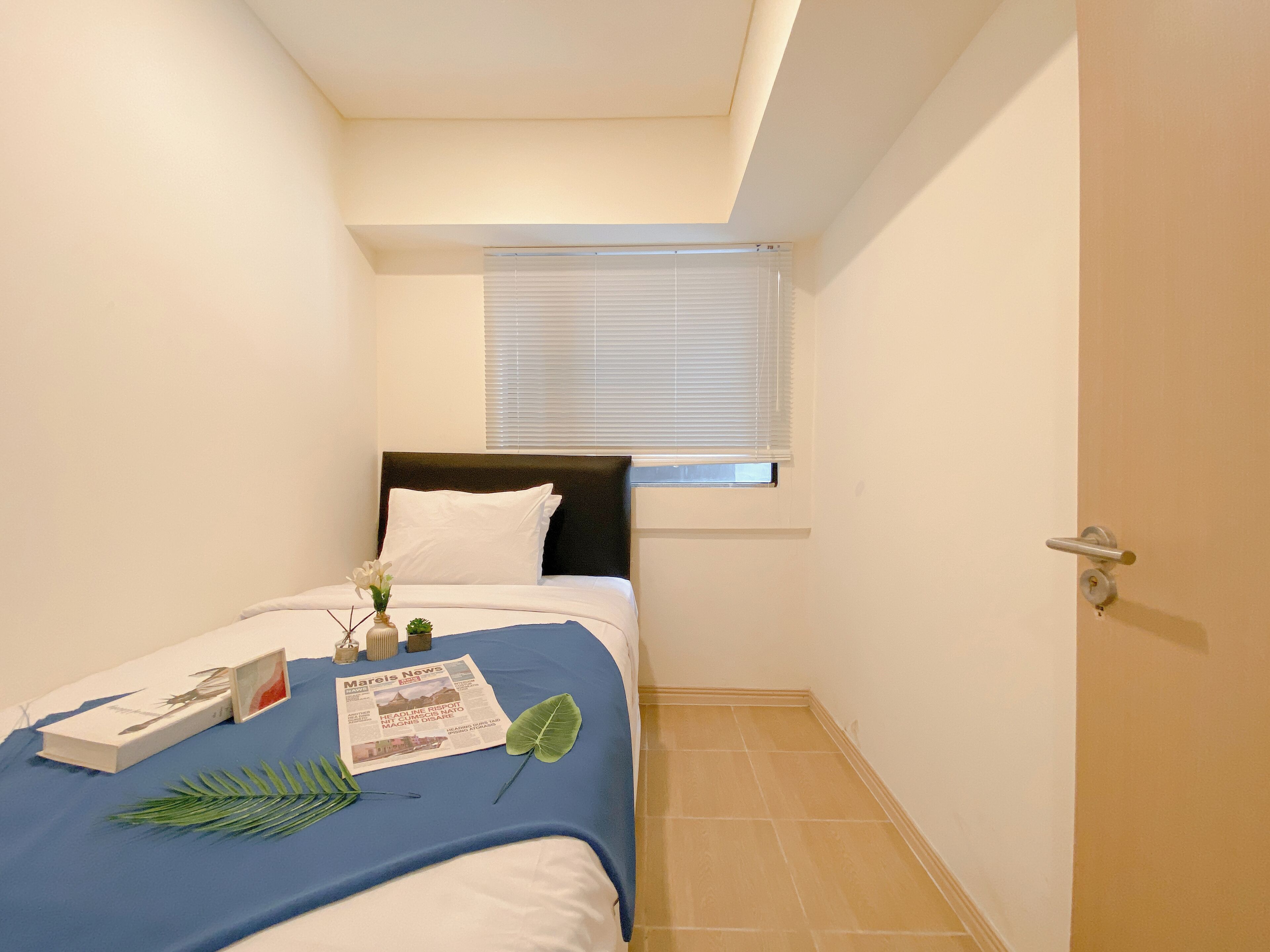 A small, neatly arranged bedroom with a single bed, a window with blinds, a door, and a tiled floor.