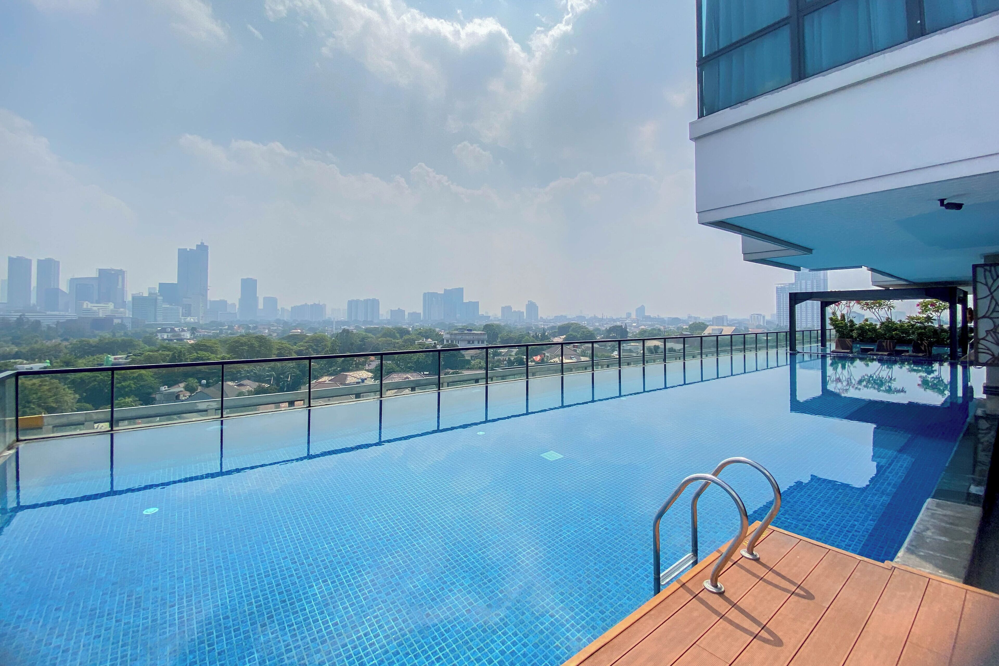 Una piscina infinita en la azotea con vistas a un horizonte urbano, una terraza de madera y una barandilla de cristal.