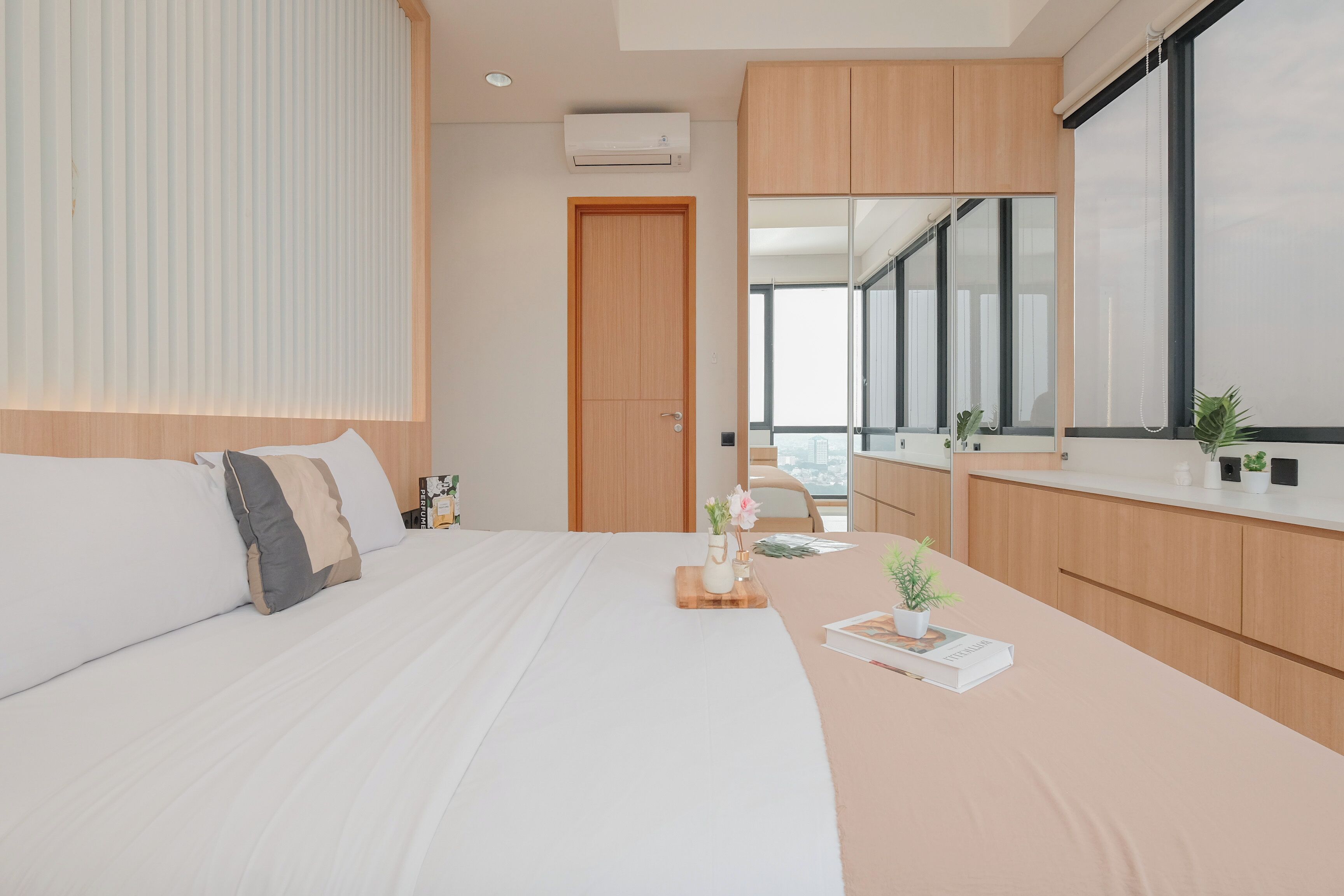 A modern bedroom with a large bed, a bedside table with a plant, a window with a view, and wooden cabinetry.