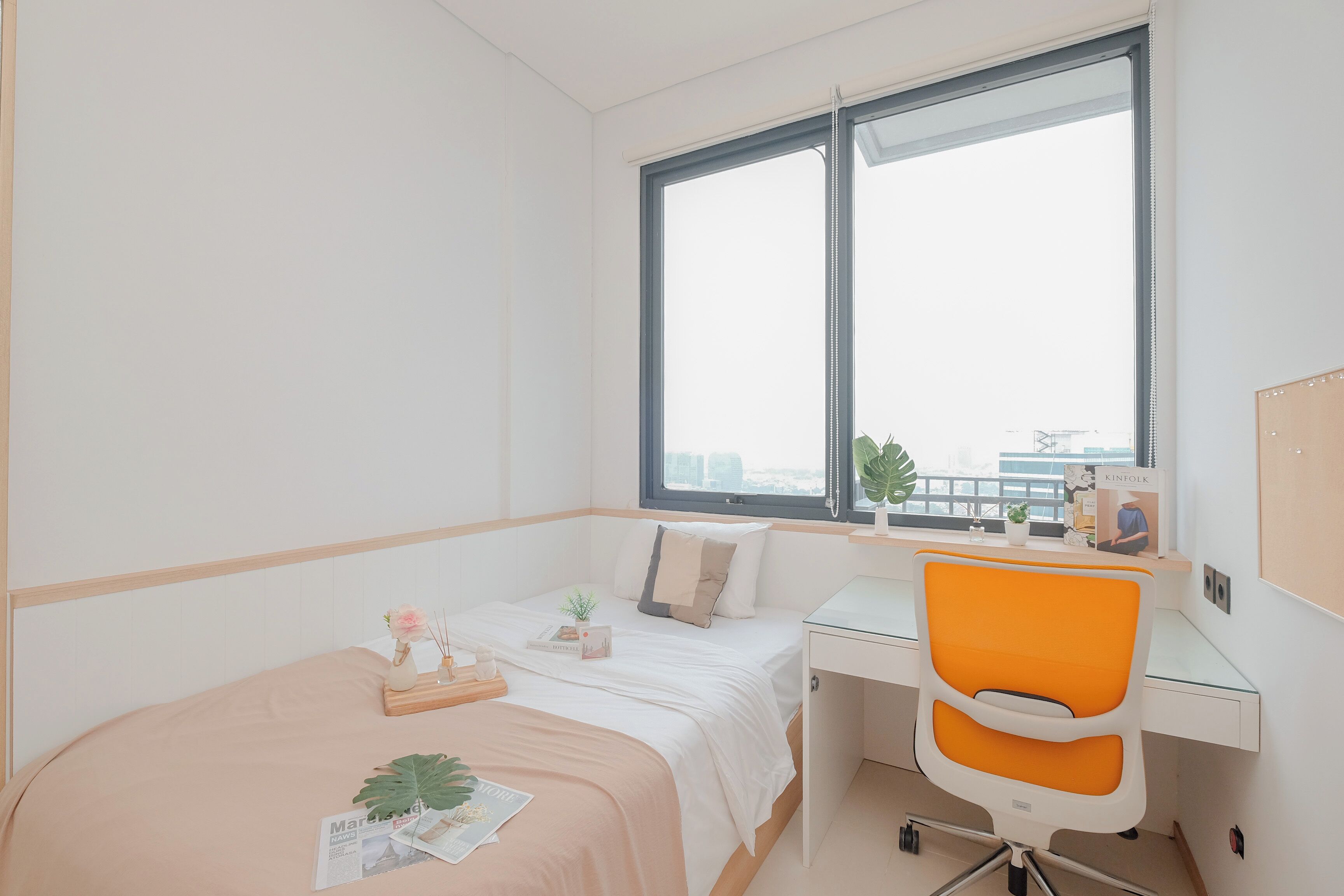 A modern bedroom with a bed, a desk, and a chair. There is a window with a view of buildings outside.