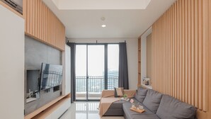 A modern living room with a grey sofa, a TV mounted on a wooden wall, a balcony with a view of buildings, and a sliding glass door. - Modern Furnished 2Br At Samara Suites Apartment (Jakarta)
