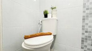 A white toilet with a brown towel on the seat, a small potted plant on top, and a tiled wall with a gray mosaic pattern. - Modern Contemporary 1Br At Tokyo Riverside Pik 2 Apartment (Teluknaga)