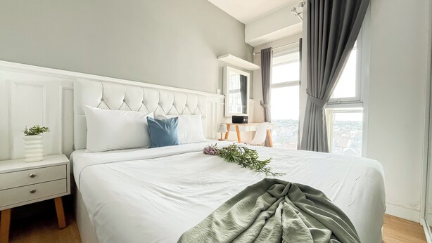 A neatly made bed with white bedding and a grey throw blanket, a bedside table with a plant, and a view of the city through large windows.