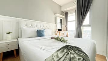 A neatly made bed with white bedding and a grey throw blanket, a bedside table with a plant, and a view of the city through large windows.
