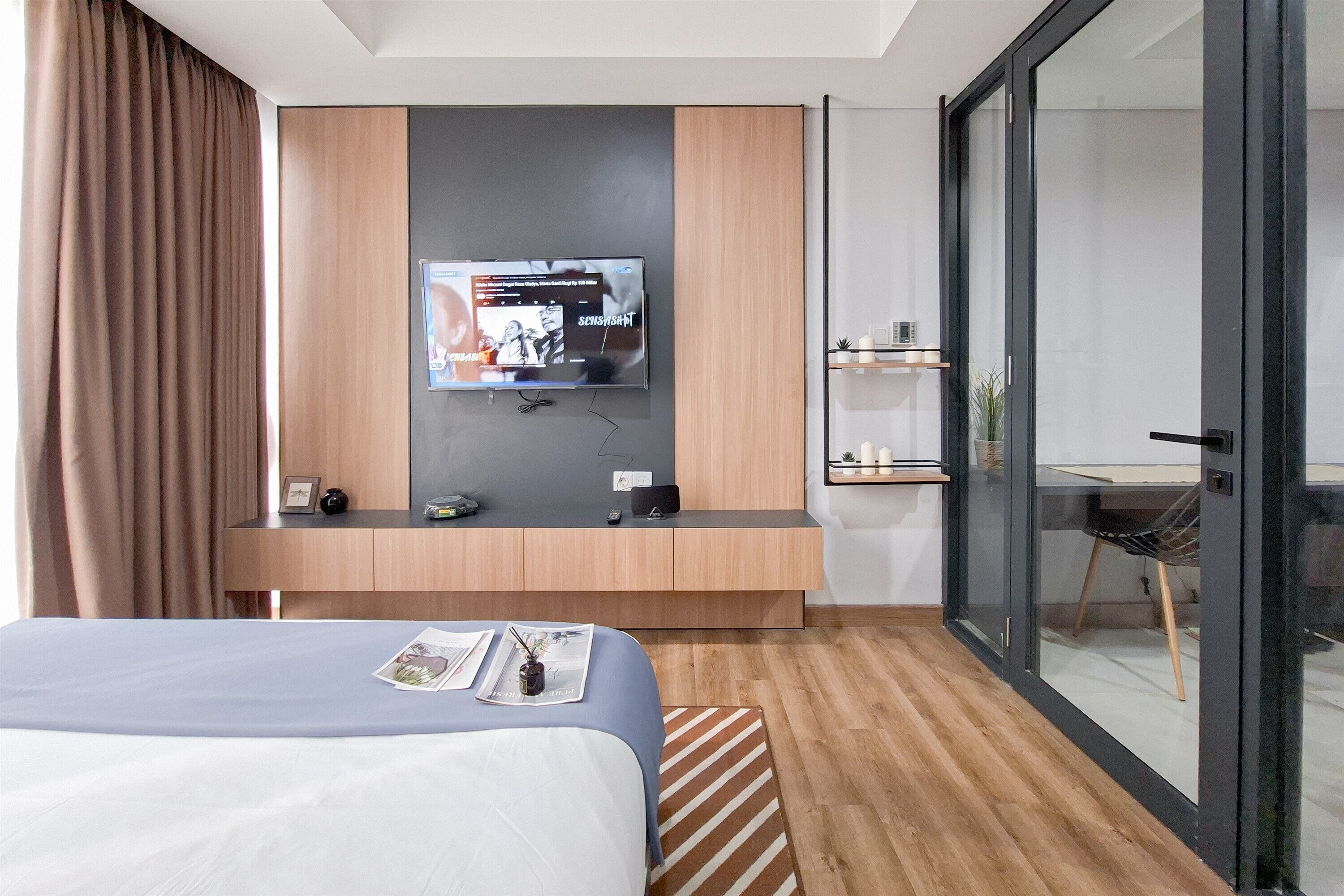 A hotel room with a bed, a TV mounted on the wall, a desk with a computer, and a sliding door leading to a balcony.