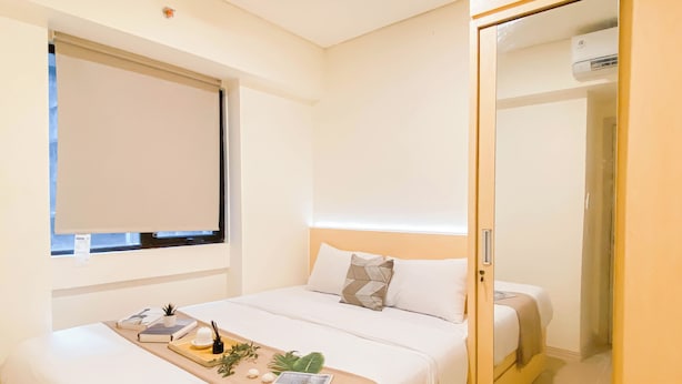 A hotel room with a bed, a bedside table with a tray, a window, and a door leading to a bathroom. - Modern Contemporary 2Br Apartment At Meikarta (Cikarang)